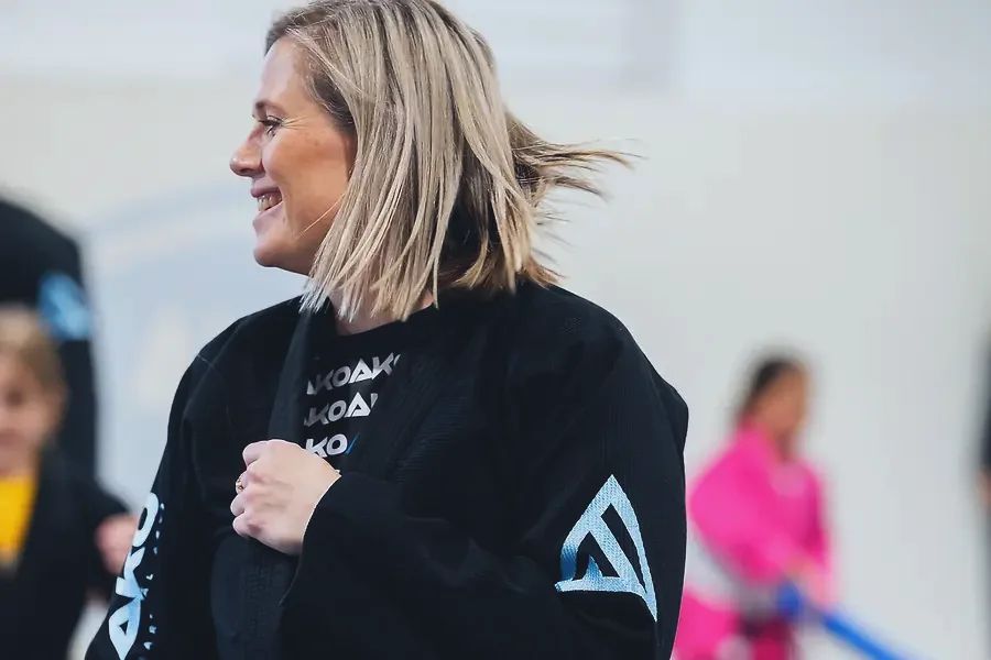 A woman tying her black belt with a young student in a pink gi behind